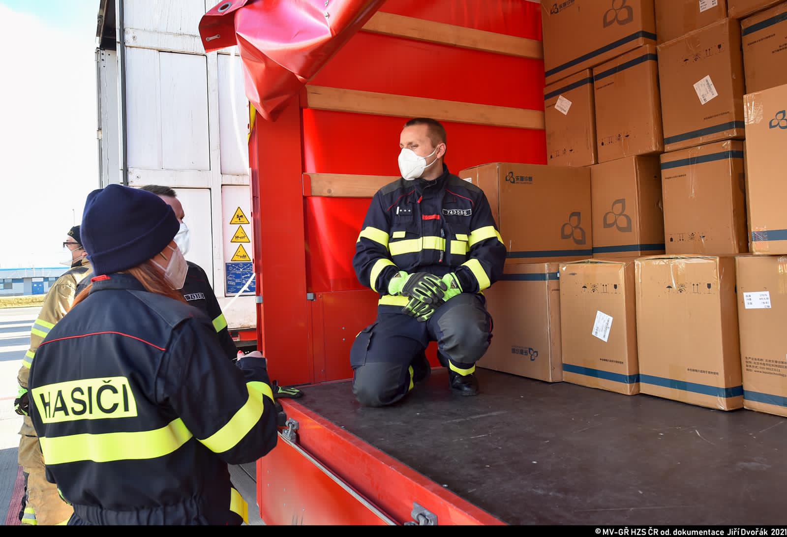 ZÚ_vykládka testů na covid na letišti Václava Havla_pohled na hasiče nakládající bedny.JPG